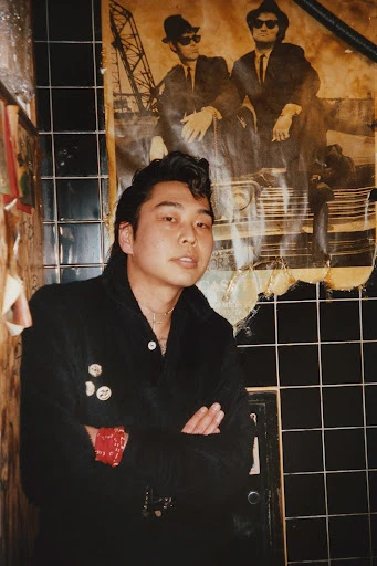 An Asian man poses indoors wearing a black jacket, with a Blues Brothers poster on the wall, featuring black and blue tiles.