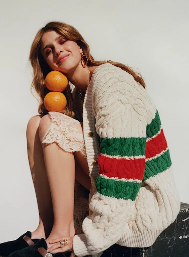 Attractive brunette woman poses in cable knit cardigan with stacked oranges, seated indoors in soft light against a plain background.