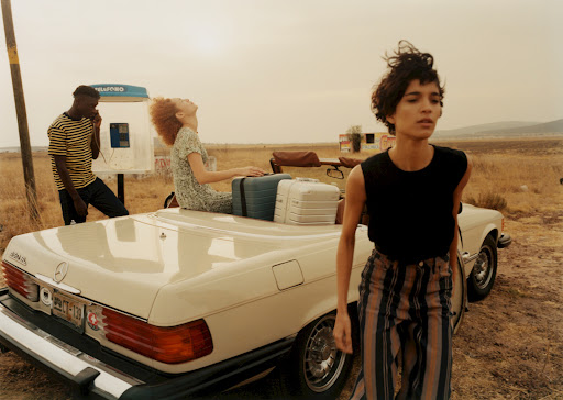 Three models pose near a vintage convertible car and an outdoor phone booth in a tan field on a cloudy day, fashion editorial.