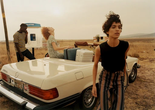 Three models pose near a vintage convertible car and an outdoor phone booth in a tan field on a cloudy day, fashion editorial.