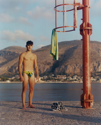 Young man in swimsuit standing on dock near water with mountain backdrop, towel hanging on red metal structure, clear blue sky.