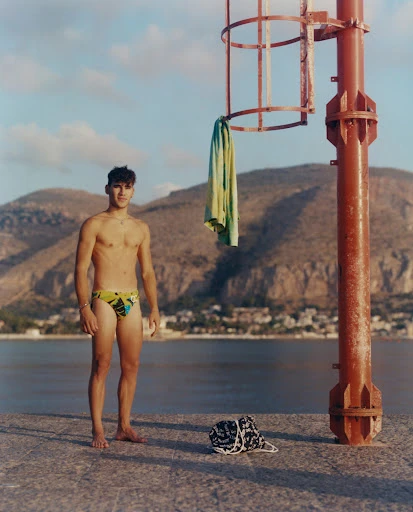 Young man in swimsuit standing on dock near water with mountain backdrop, towel hanging on red metal structure, clear blue sky.