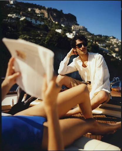 Two people relaxing on a boat on a sunny day. One is reading a newspaper while the other is looking at the coastal landscape.