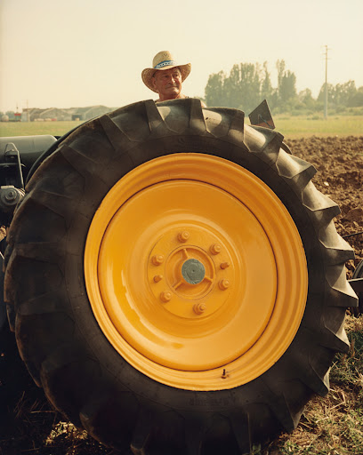 An adult male wearing a hat sits on a tractor with a very large yellow and black tire in a field with a farm in the background.