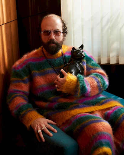 Bearded adult man wearing a colorful striped outfit and glasses poses with a black cat sculpture indoors against a wood-paneled wall.