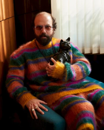 Bearded adult man wearing a colorful striped outfit and glasses poses with a black cat sculpture indoors against a wood-paneled wall.