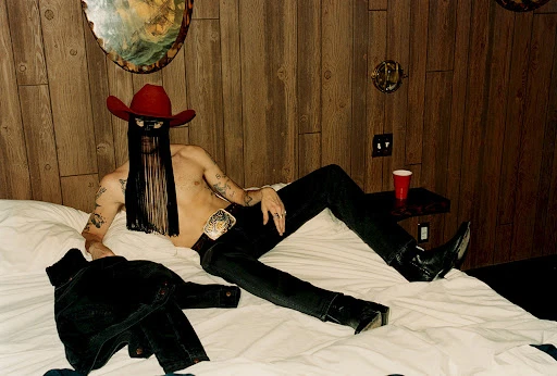 Man in cowboy hat and fringed mask lies on a bed in a wood-paneled room, wearing jeans and boots, with a denim jacket on the bed.