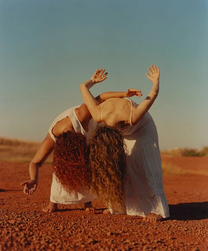 Two women in white dresses are close together in a dance pose on red earth under a blue sky, showing off their curly hair.