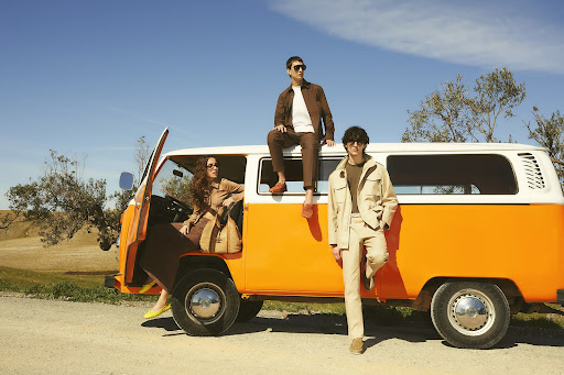 Three stylish adults pose with a vintage orange and white camper van under a bright blue sky, embodying travel and fashion.