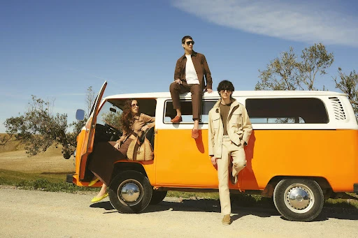 Three stylish adults pose with a vintage orange and white camper van under a bright blue sky, embodying travel and fashion.