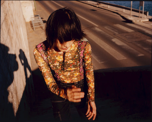 A woman in a floral top stands on a city street with her shadow cast on a concrete wall during daytime, captured in a medium shot.