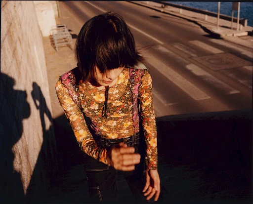 A woman in a floral top stands on a city street with her shadow cast on a concrete wall during daytime, captured in a medium shot.