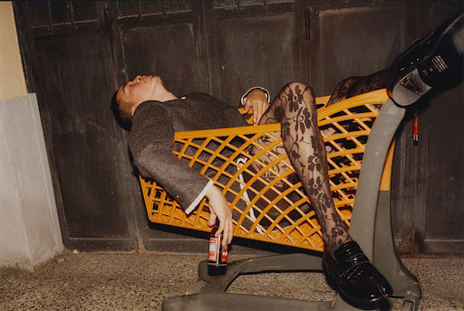 A man in formal wear and floral tights reclines in a shopping cart, holding a beverage outdoors with a dark, moody aesthetic.