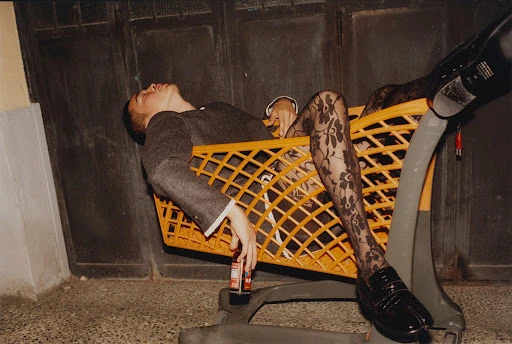 A man in formal wear and floral tights reclines in a shopping cart, holding a beverage outdoors with a dark, moody aesthetic.