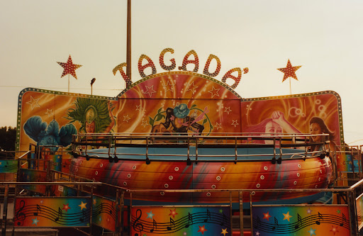Carousel ride at a carnival, featuring a multi-colored spinning platform with decorative artwork, stars, and musical notes against a light sky.