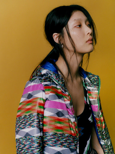 Portrait of an Asian woman in a patterned jacket against a yellow backdrop. She has dark hair pulled back and is looking off to the side.