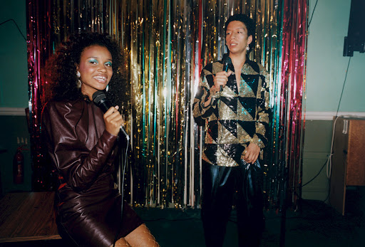Two singers, a woman and a non-binary person, perform on stage with microphones in a dimly lit room with tinsel backdrop.