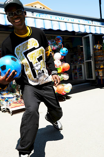 Smiling African American man in black clothing and sunglasses stands on a paved surface near a building with balls and text.