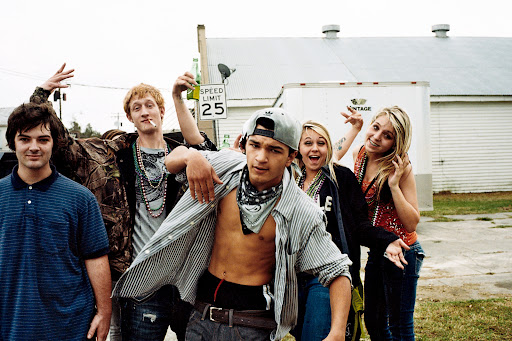 Group of six young adults posing outdoors, wearing casual clothing and jewelry, with a white building in the background during the daytime.