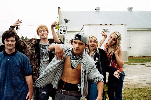 Group of six young adults posing outdoors, wearing casual clothing and jewelry, with a white building in the background during the daytime.