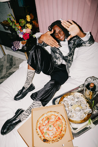 Person poses on bed with pizza and oysters. Model wears black blazer, white shirt, zebra socks and loafers in indoor studio.