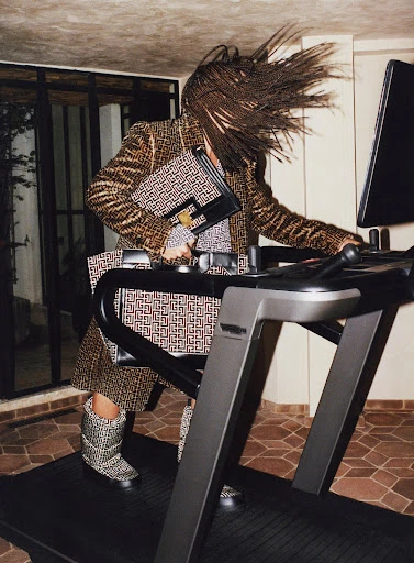 Fashion shot of a woman on a treadmill wearing a patterned coat, skirt, and boots, holding patterned bags. Indoor setting.