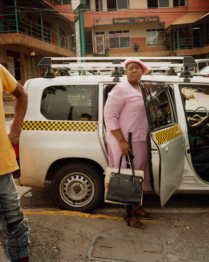 Mature woman in a pink suit exiting a white taxi in an urban setting, carrying a black purse and a walking cane. 