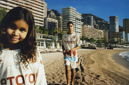 A brother and sister pose on a sandy beach in Monaco, France, with blue sky, city buildings, and mountains in the background on a sunny day.