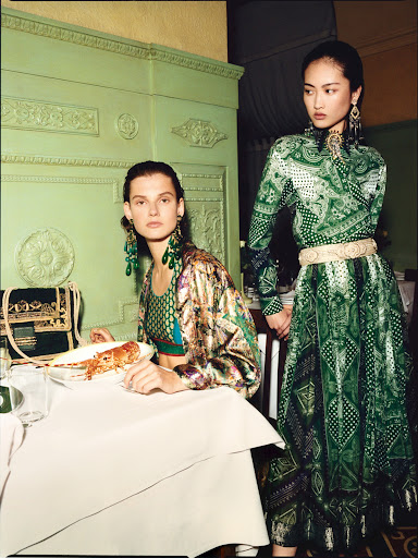 Two women in patterned dresses pose at a table with a plate of food in a staged fashion photograph, indoors, with a light green wall.