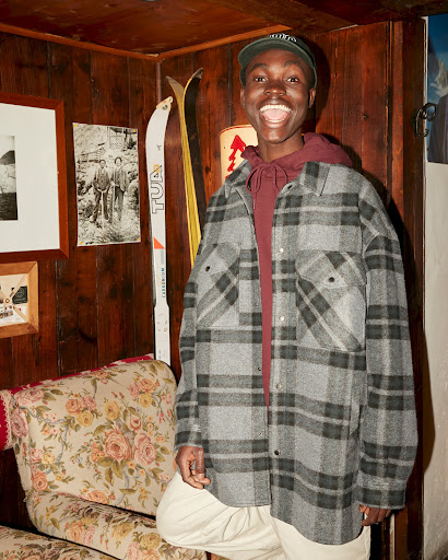 Smiling African American man in a grey and black plaid jacket, maroon hoodie, and green baseball cap stands indoors near a floral chair.