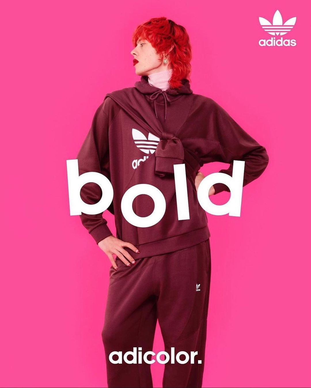 Model in a maroon Adidas tracksuit and matching sweatshirt poses against a bright pink background for a fashion brand ad.