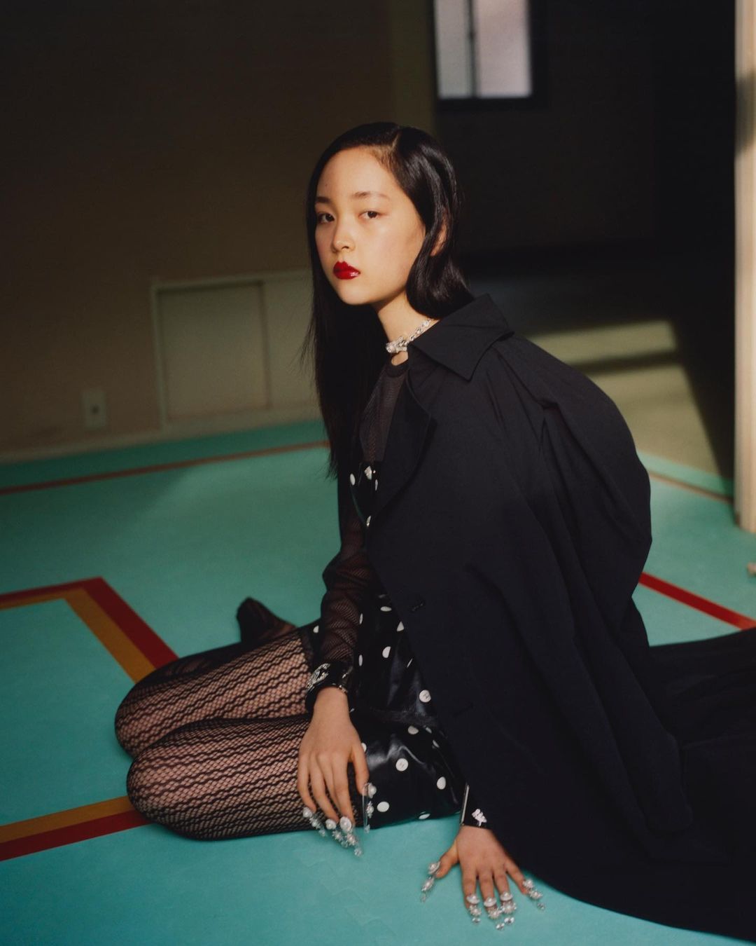 An Asian woman is posing on a turquoise floor wearing a black dress with polka dots, black fishnet tights, and a long black coat.