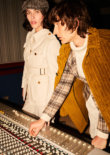 Two adult models, a male and a female, pose in a recording studio with a mixing console. The lighting is studio with warm tones.