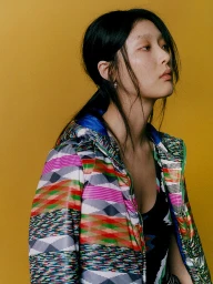 Portrait of an East Asian woman wearing a colourful geometric patterned puffer jacket against a solid yellow background.