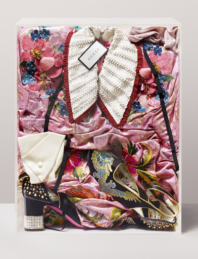 Still life of a pink floral embroidered Gucci dress, white frilled collar, platform heels, and a clutch in a clear display case.