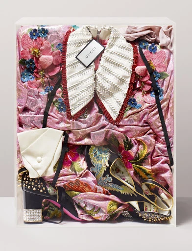 Still life of a pink floral embroidered Gucci dress, white frilled collar, platform heels, and a clutch in a clear display case.