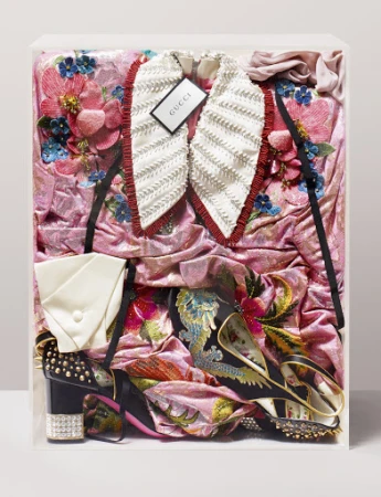 Still life of a pink floral embroidered Gucci dress, white frilled collar, platform heels, and a clutch in a clear display case.