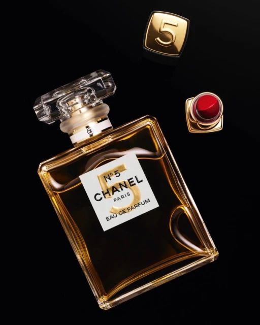 Elevated view of Chanel No. 5 perfume bottle, red lipstick, and gold Chanel cap on a black surface. Luxury beauty and cosmetics product photography.