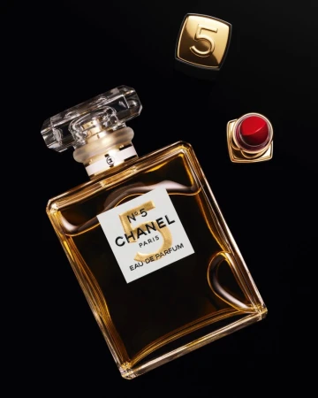 Elevated view of Chanel No. 5 perfume bottle, red lipstick, and gold Chanel cap on a black surface. Luxury beauty and cosmetics product photography.