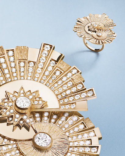 Close-up of a gold statement ring and jewelry piece adorned with diamond-like stones against a soft blue backdrop in a studio setting.