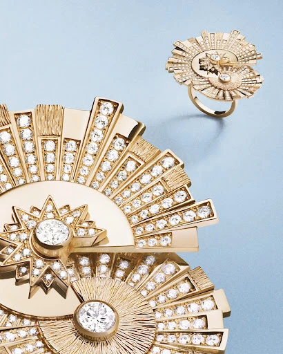 Close-up of a gold statement ring and jewelry piece adorned with diamond-like stones against a soft blue backdrop in a studio setting.