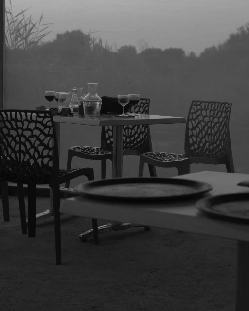 Black and white shot of an outdoor dining area with prepared tables, featuring glassware, carafe, and modern lattice-back chairs.