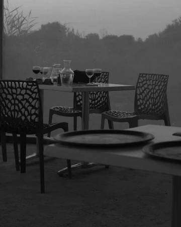 Black and white shot of an outdoor dining area with prepared tables, featuring glassware, carafe, and modern lattice-back chairs.