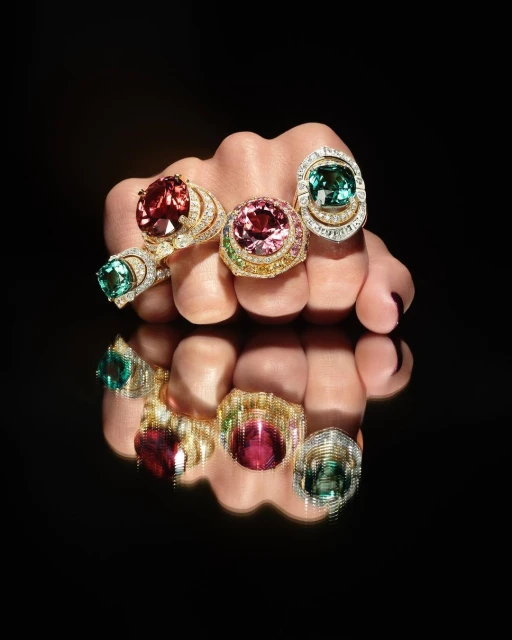 Close-up of a hand adorned with several large, colorful gemstone rings with diamond halos, reflected on a glossy black surface.