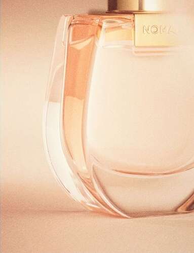 Close-up of a clear glass perfume bottle with a gold label, on a soft peach-colored background, showcasing its elegant design.