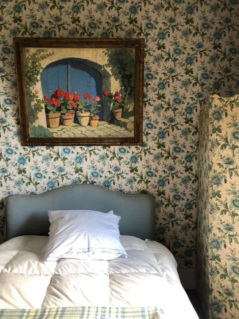 Bedroom interior featuring a bed with blue floral wallpaper and an framed painting above the headboard with flower pots. 