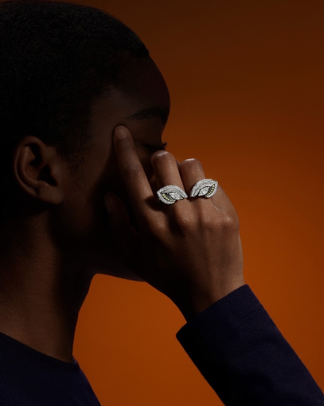 Close-up of a dark-skinned adult female model wearing two leaf-shaped diamond rings against a deep orange background.