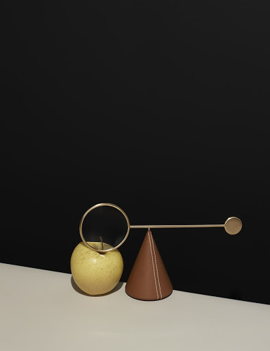 Still life shot of an apple with a gold ring on top, a cone, and a rod against a black background.