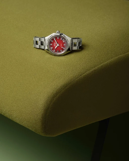 Wristwatch with a stainless steel bracelet on a green textured surface, showcasing a red dial and silver hands in a product shot.