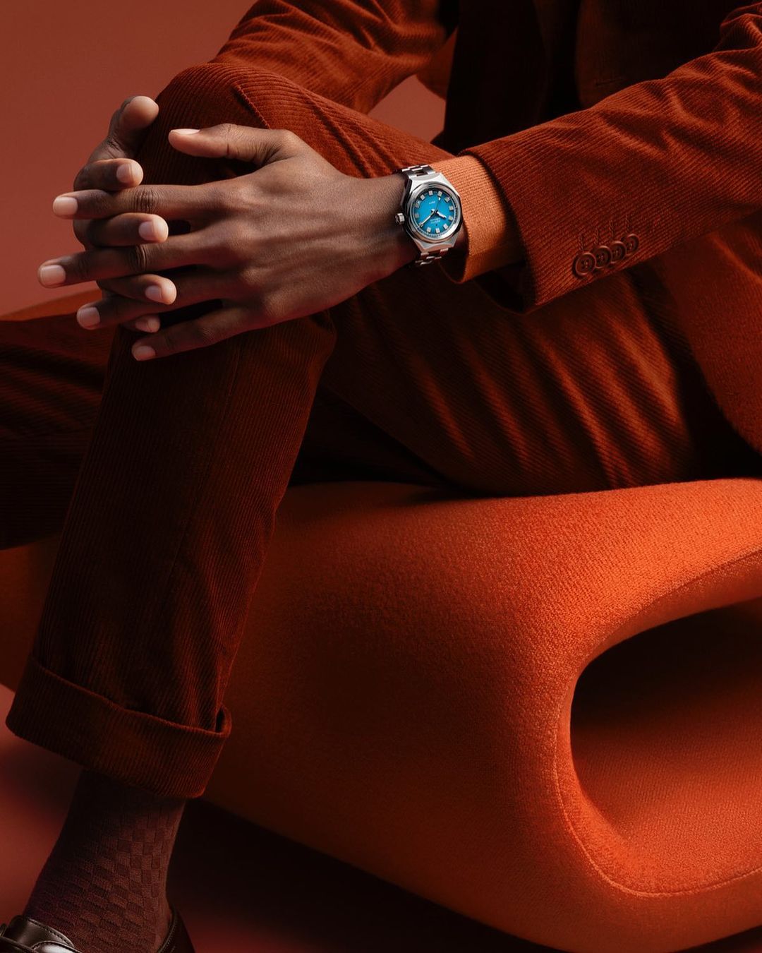A person wearing a brown corduroy suit and silver watch with a blue face sits on an orange chair against a warm, solid-color background.